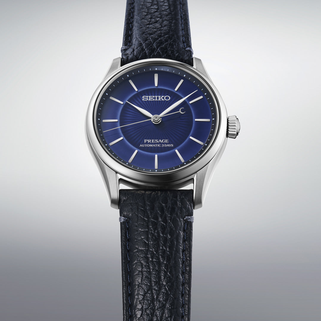The Seiko Presage Classic Series 145th Anniversary Automatic 39.6mm Watch Limited Edition of 1,500 Pieces features a striking blue dial with silver hour markers and a black textured leather strap, all set against a subtle gradient backdrop. This limited-edition timepiece by Seiko beautifully combines refined style with timeless reliability.
