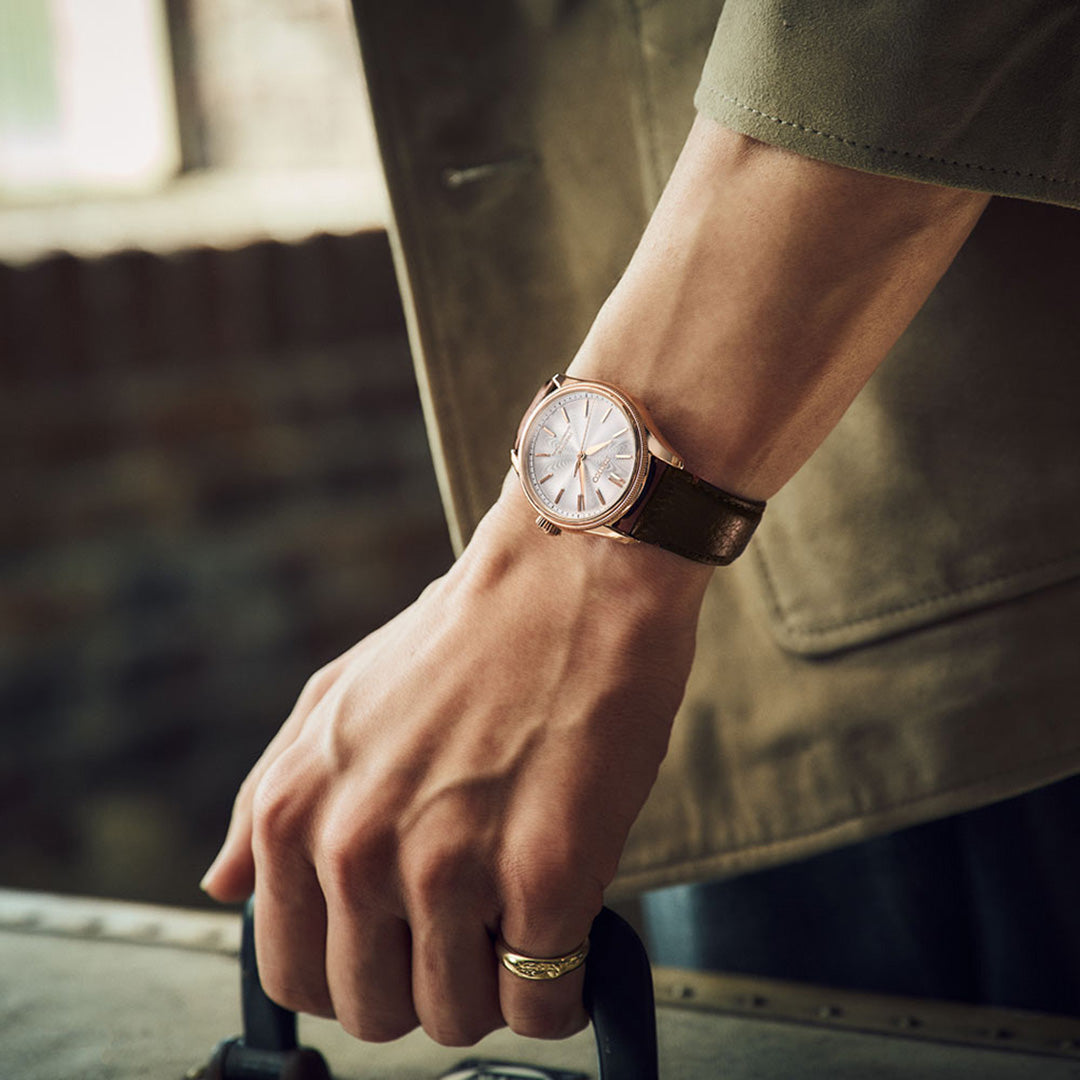 A close-up of a person’s hand holding a suitcase handle, wearing the Seiko Presage Classic Series Tomioka Silk Promotion Organization Automatic 38mm Watch Limited Edition of 2,000 pieces. The elegant timepiece by Seiko, featuring a brown leather strap and gold ring, perfectly complements the person’s olive green jacket for a refined travel look.
