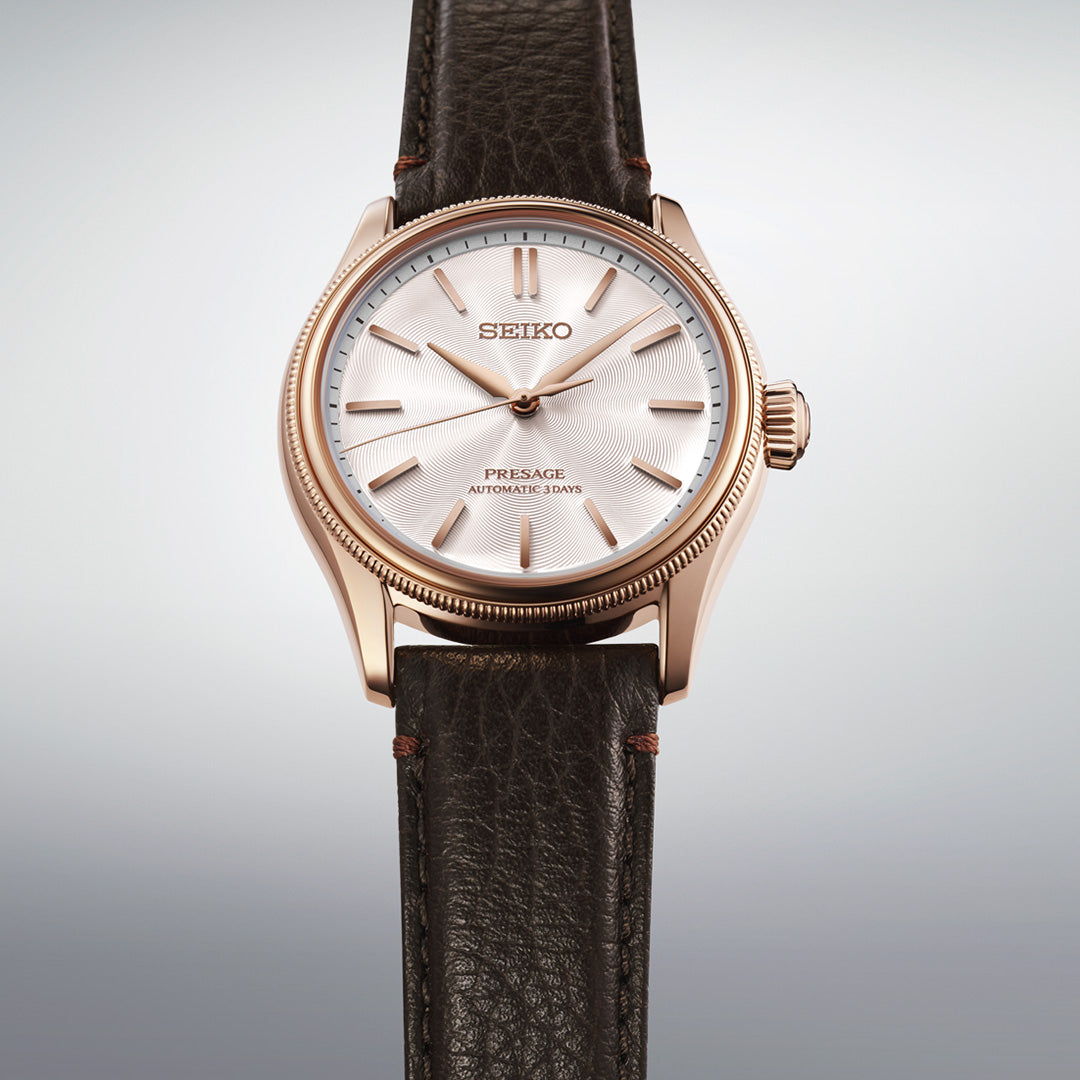 A Seiko Presage Classic Series Tomioka Silk Promotion Organization Automatic 38mm Limited Edition watch (one of 2,000 pieces), featuring a white textured dial with gold hour markers and case, and a dark brown leather strap, is displayed against a plain light grey background.
