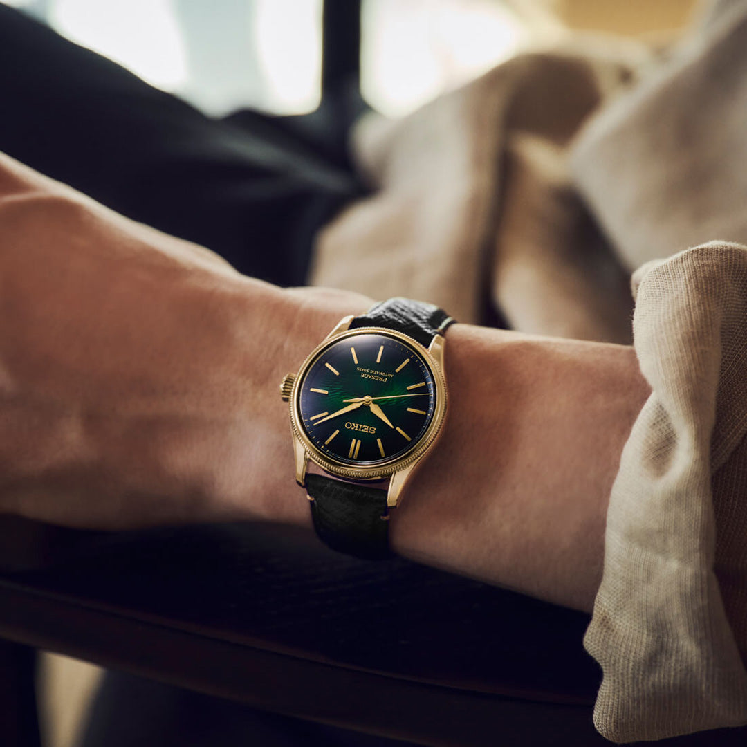 A person wearing the Seiko Presage Classic Series Tradman’s Bonsai Automatic 38mm Watch, a limited edition of 2,000 pieces by Seiko, featuring a gold case, black leather strap, and dark green dial, rests their arm on a chair.
