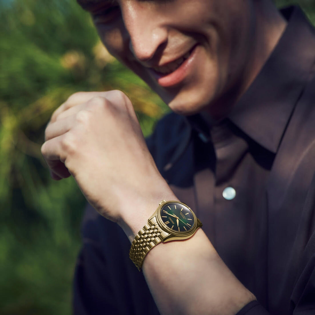 A person wearing a dark shirt smiles and looks down at their Seiko Presage Classic Series Tradman’s Bonsai Automatic 38mm Watch, a limited edition of 2,000 pieces by Seiko, while standing outdoors with green foliage softly blurred in the background.
