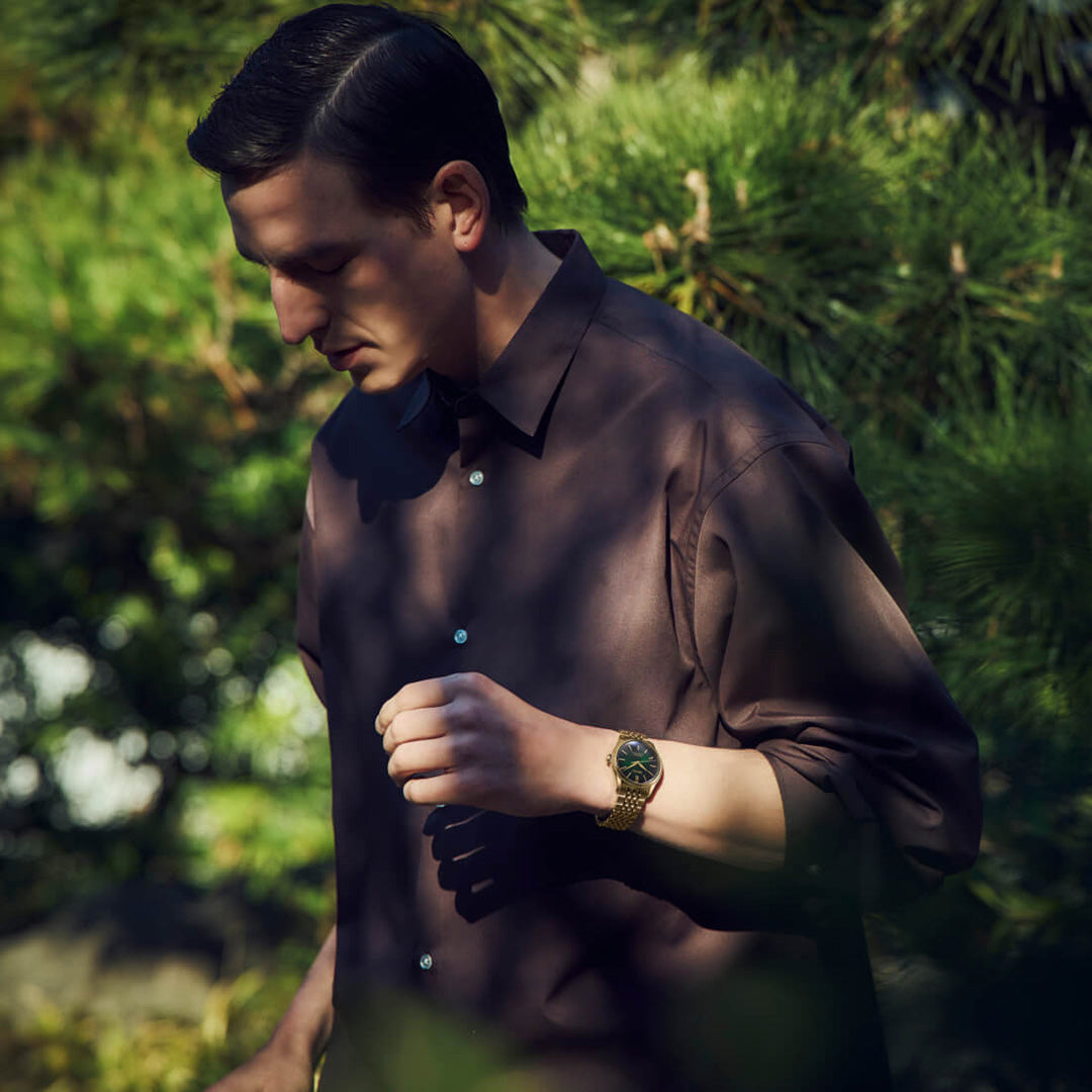 A man in a brown shirt stands outdoors amid lush green foliage, gazing down thoughtfully. On his wrist, he wears the Seiko Presage Classic Series Tradman’s Bonsai Automatic 38mm Watch—a limited edition of only 2,000 pieces—its gold accents gleaming where sunlight breaks through the shadows.