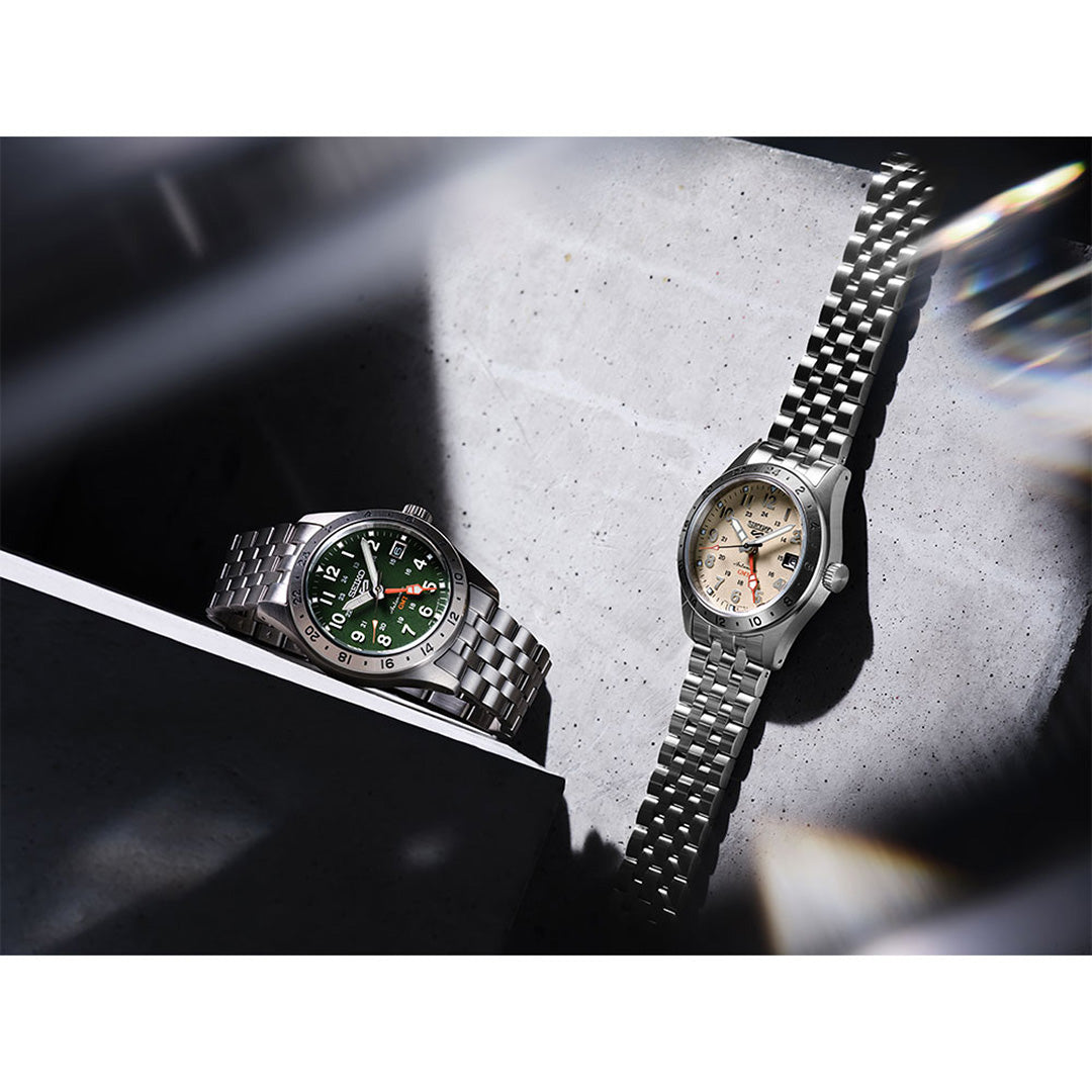 Two stainless steel wristwatches, both Seiko 5 Sports Field Series GMT Automatic 39.3mm models—one with a green face and the other with a silver face—rest on a concrete surface, illuminated by dramatic lighting and softened shadows.