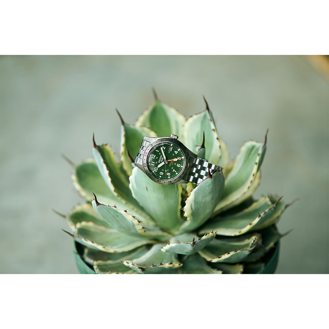 A Seiko 5 Sports Field Series GMT Automatic 39.3mm Watch by Seiko, featuring a green dial and a stainless steel case, rests atop a spiky green succulent plant, its sleek silver finish standing out elegantly against the softly blurred gray background.