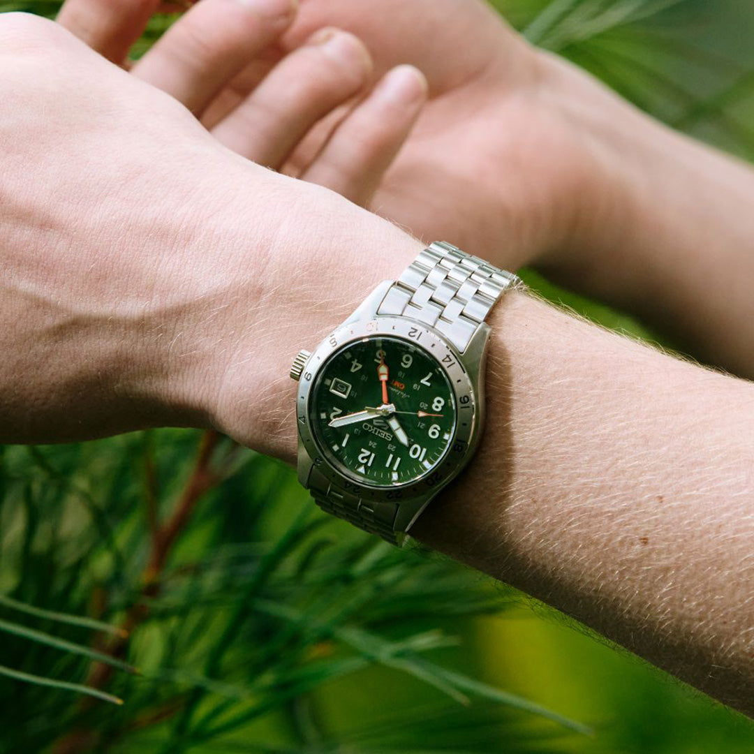 A person’s wrist wearing a Seiko 5 Sports Field Series GMT Automatic 39.3mm stainless steel watch by Seiko, featuring a green dial with white numerals, set against a blurred backdrop of greenery.