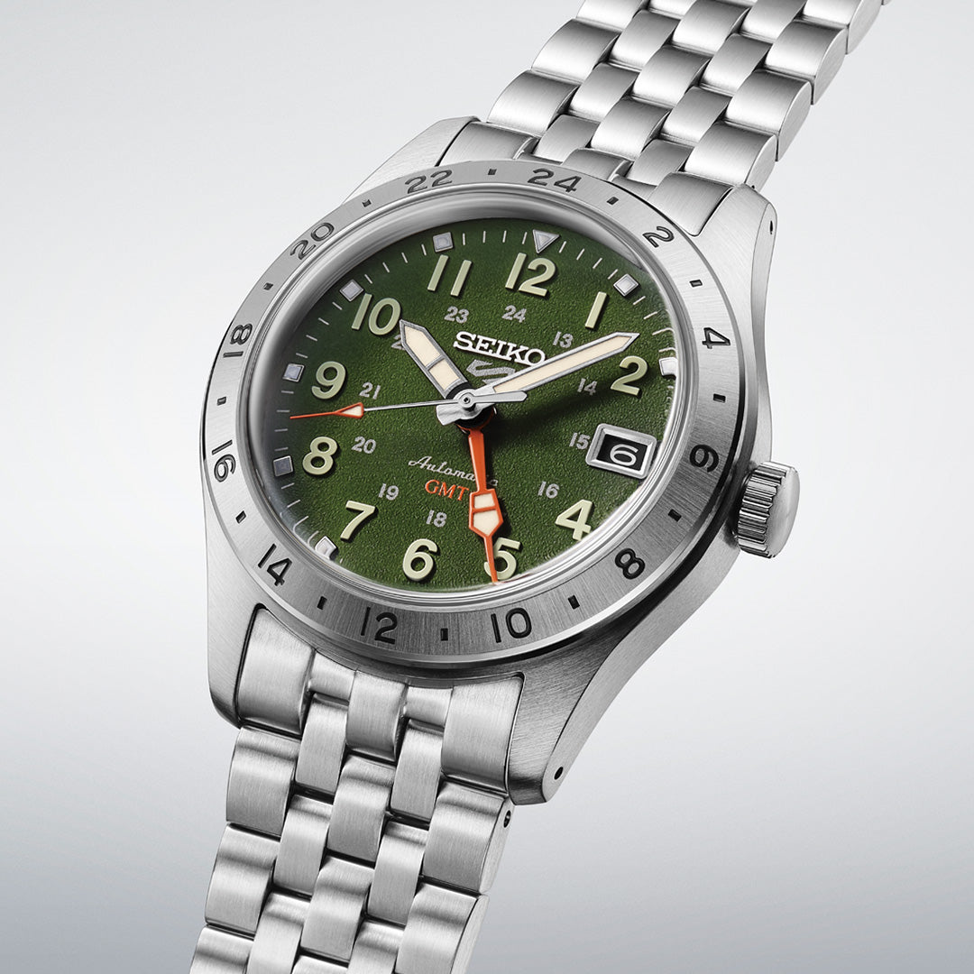 A Seiko 5 Sports Field Series GMT Automatic 39.3mm watch by Seiko, featuring a green dial, silver stainless steel bezel, metal bracelet, white hour markers, a date window at 3 o’clock, and an orange GMT hand.