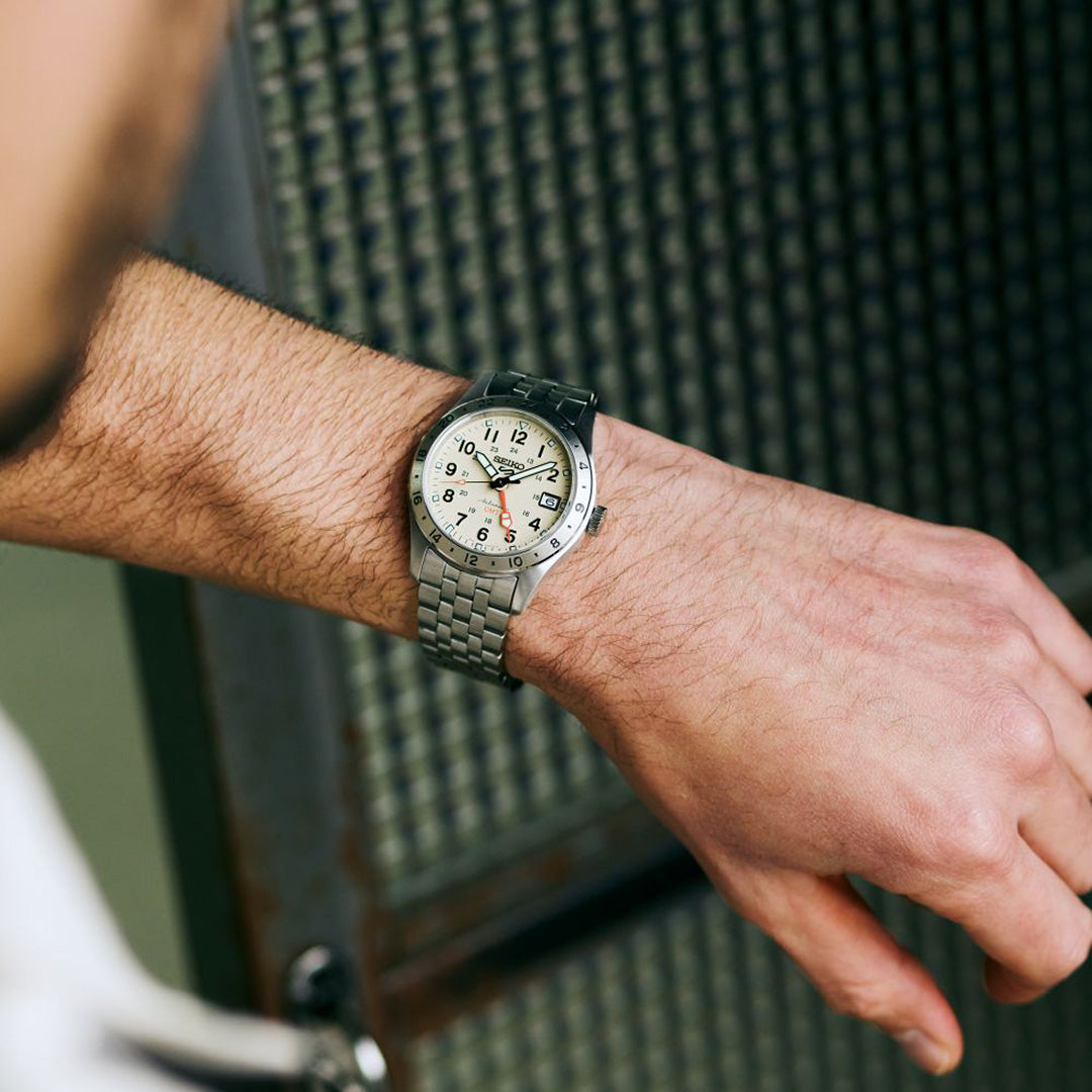 A person wearing a Seiko 5 Sports Field Series GMT Automatic 39.3mm Watch by Seiko stands in front of a metal, grid-patterned background.