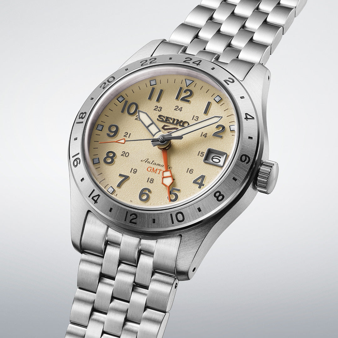 Introducing the Seiko 5 Sports Field Series GMT Automatic 39.3mm Watch by Seiko, featuring a stainless steel case and bracelet, a beige dial with a date display at the 3 o’clock position, and a distinctive red GMT hand. The bezel showcases 24-hour markings, adding to the functionality of this automatic timepiece.