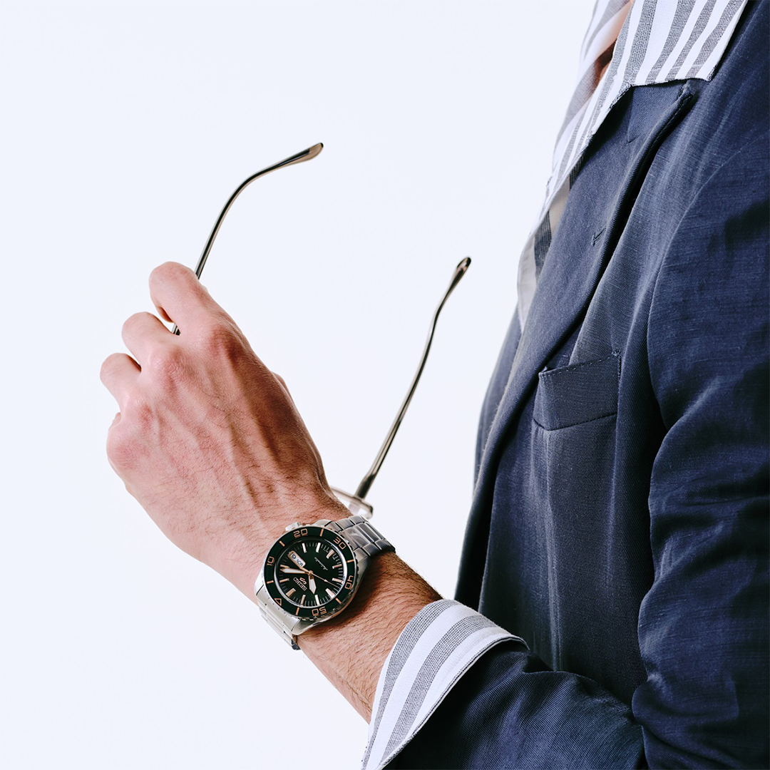 A person in a blue suit jacket and striped shirt holds eyeglasses while wearing a Seiko 5 Sports SKX Series Automatic 42.5mm Watch with a black dial and a rotating bezel.