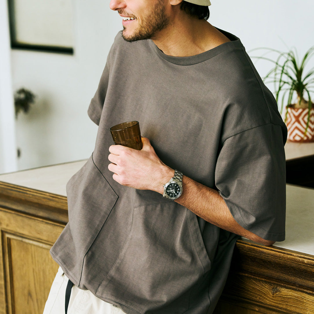A man in a loose gray t-shirt and beige cap leans on a counter, smiling and holding a wooden cup. He is wearing the Seiko 5 Sports Field Series Automatic 41mm Watch by Seiko, which features a metal bracelet, and is standing beside a plant in a patterned pot.