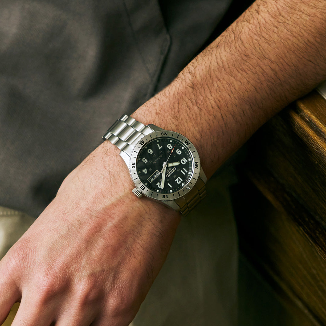 A person wears a Seiko 5 Sports Field Series Automatic 41mm Watch by Seiko, with a silver metal bracelet and black dial featuring LumiBrite hands, on their left wrist as they rest their hand on a wooden surface.