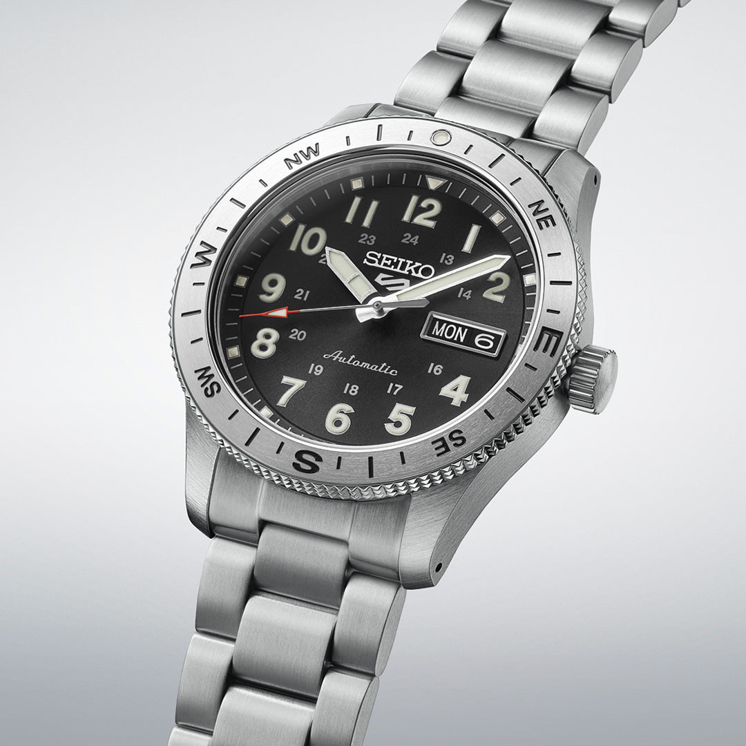 The Seiko 5 Sports Field Series Automatic 41mm Watch by Seiko features a silver metal bracelet band, a black dial with prominent white LumiBrite-coated numerals for improved visibility, and a day-date window, displayed against a light gray background.