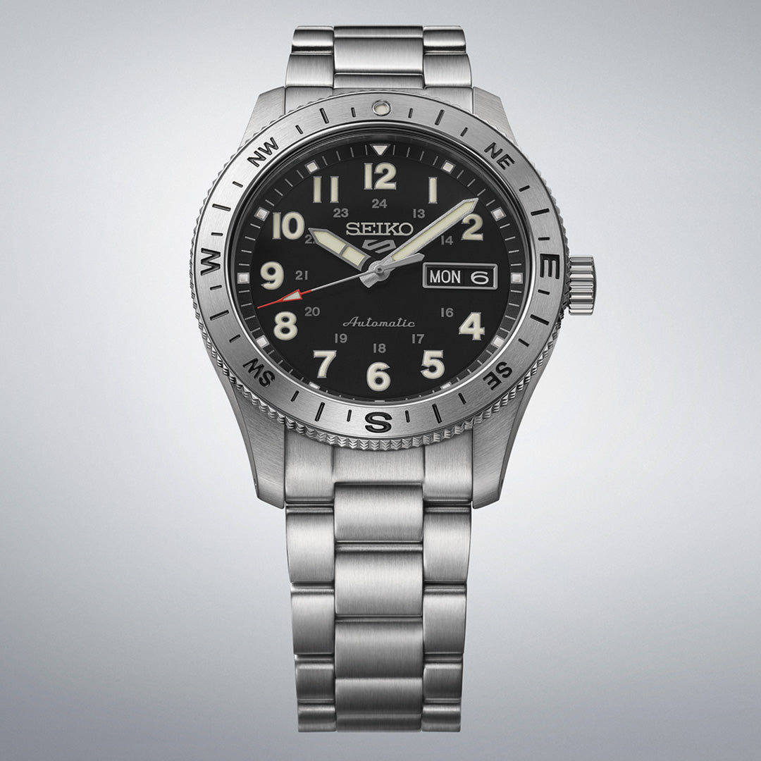 The Seiko 5 Sports Field Series Automatic 41mm Watch by Seiko features a stainless steel bracelet, black dial with LumiBrite hands and hour markers, a silver bezel marked with compass directions, and a day-date window at 3 o’clock, displayed against a plain gray background.