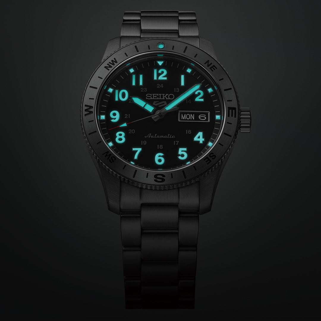 The Seiko 5 Sports Field Series Automatic 41mm Watch by Seiko features a black design with a durable metal bracelet. Its LumiBrite technology makes the numbers, hour markers, and hands glow teal in the dark. The watch displays a day-date window showing “MON 6,” and compass markings are engraved on the bezel.