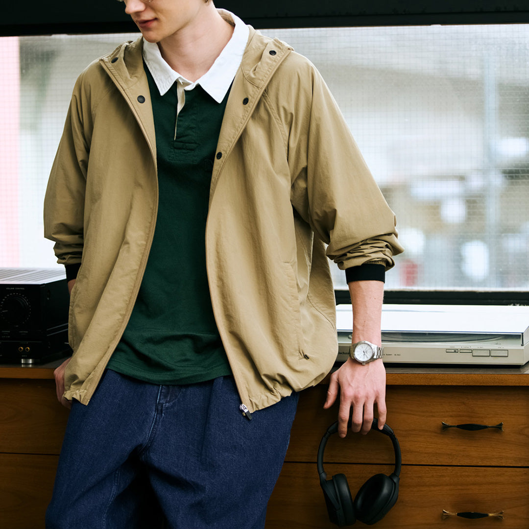 A person wearing a tan jacket, green shirt, and blue pants leans against a wooden desk with a Seiko 5 Sports Field Series Automatic 41mm Watch by Seiko on their wrist. The metal bracelet of the watch catches the light as their arm rests nearby, while headphones and audio equipment are neatly arranged on the desk by the window.