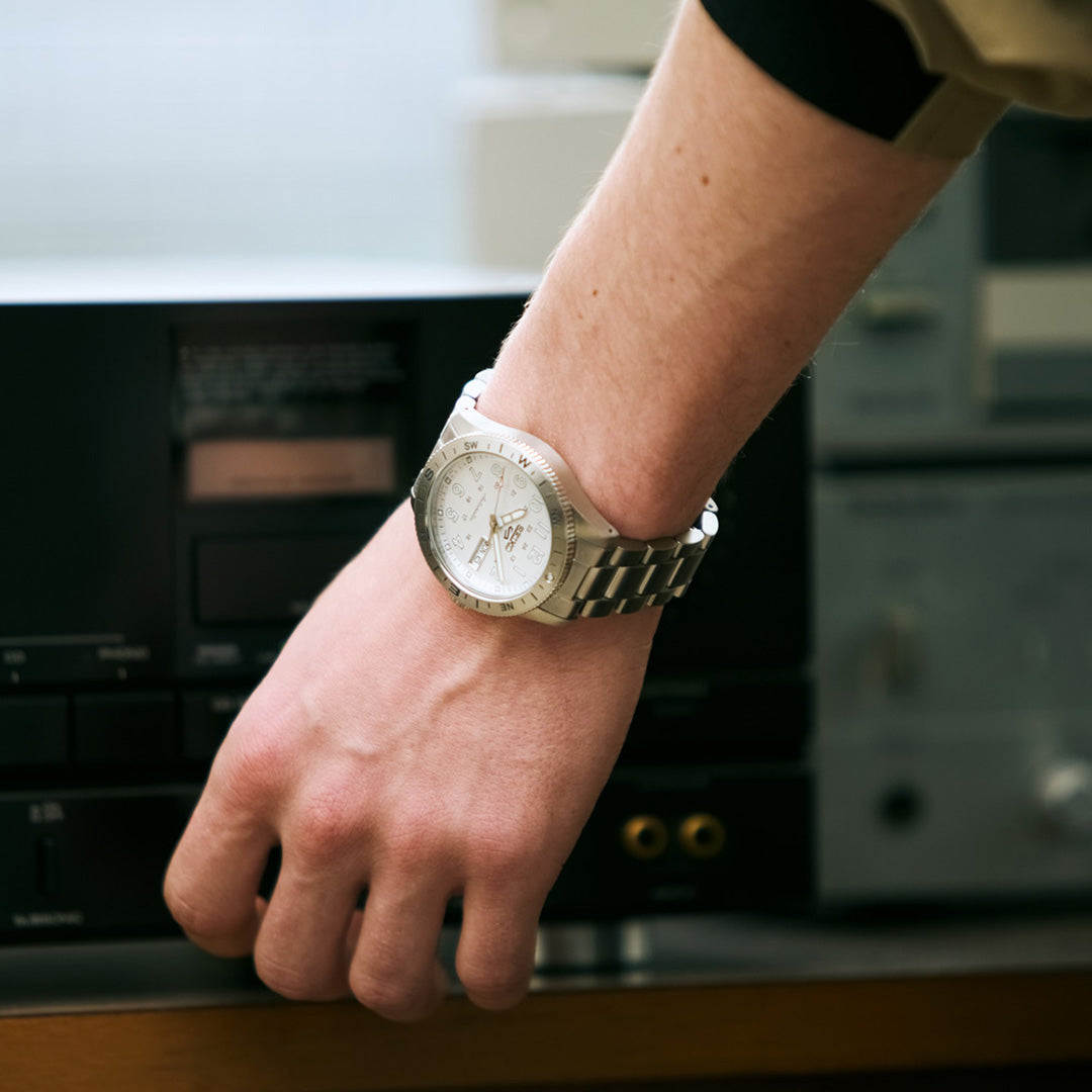 A person wearing a Seiko 5 Sports Field Series Automatic 41mm watch by Seiko with a metal bracelet is adjusting an audio system or stereo component with their right hand.