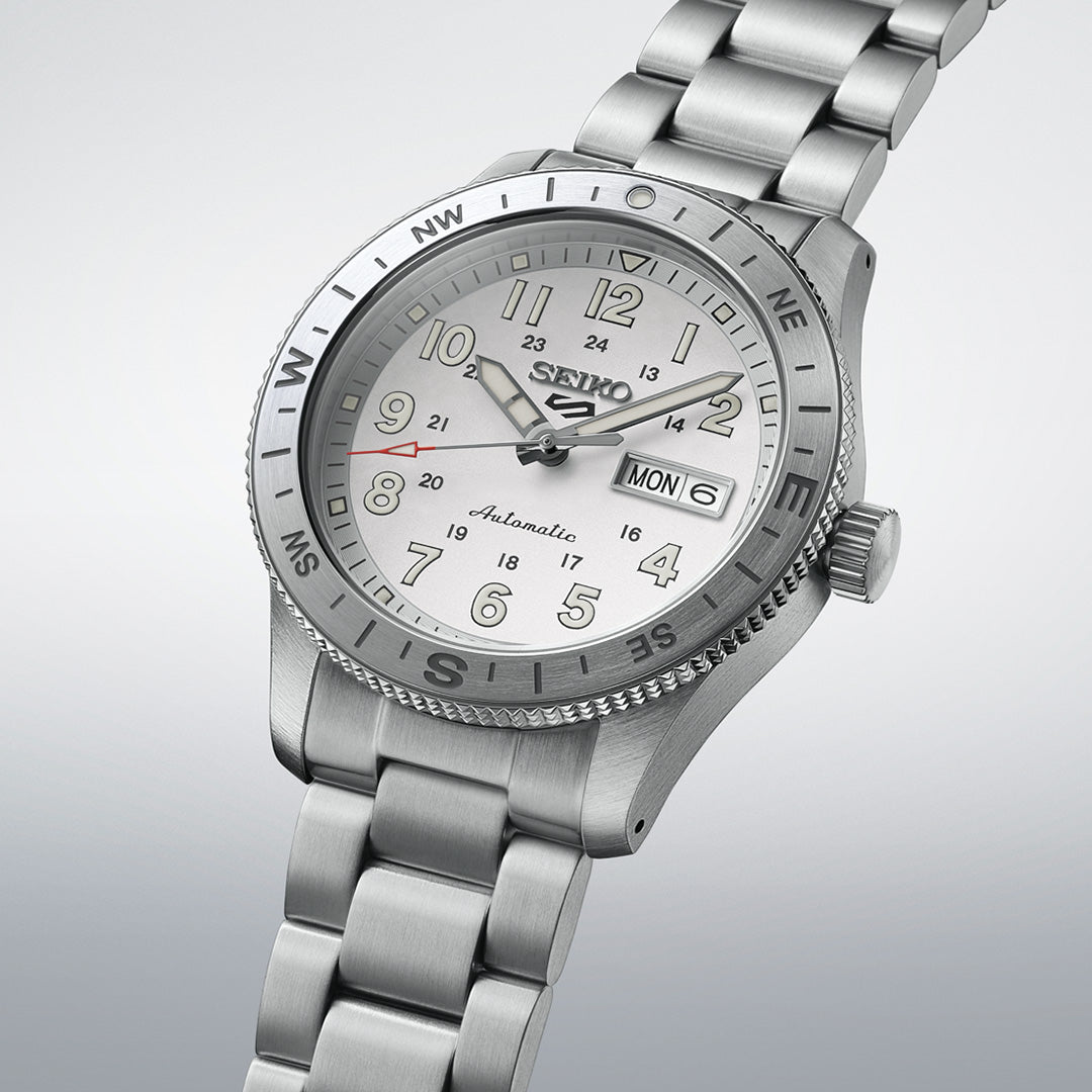 A Seiko 5 Sports Field Series Automatic 41mm Watch by Seiko, featuring a stainless steel case, silver dial, compass bezel, LumiBrite hands and markers, day and date display, and a metal bracelet, displayed against a light gradient background.