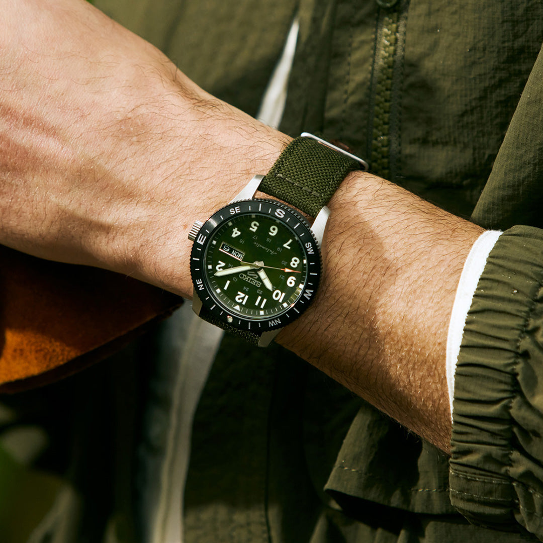 A person in an olive green jacket displays the Seiko 5 Sports Field Series Automatic 41mm Watch by Seiko on their left wrist, featuring a green dial with white LumiBrite-enhanced numerals and a durable matching nylon strap.