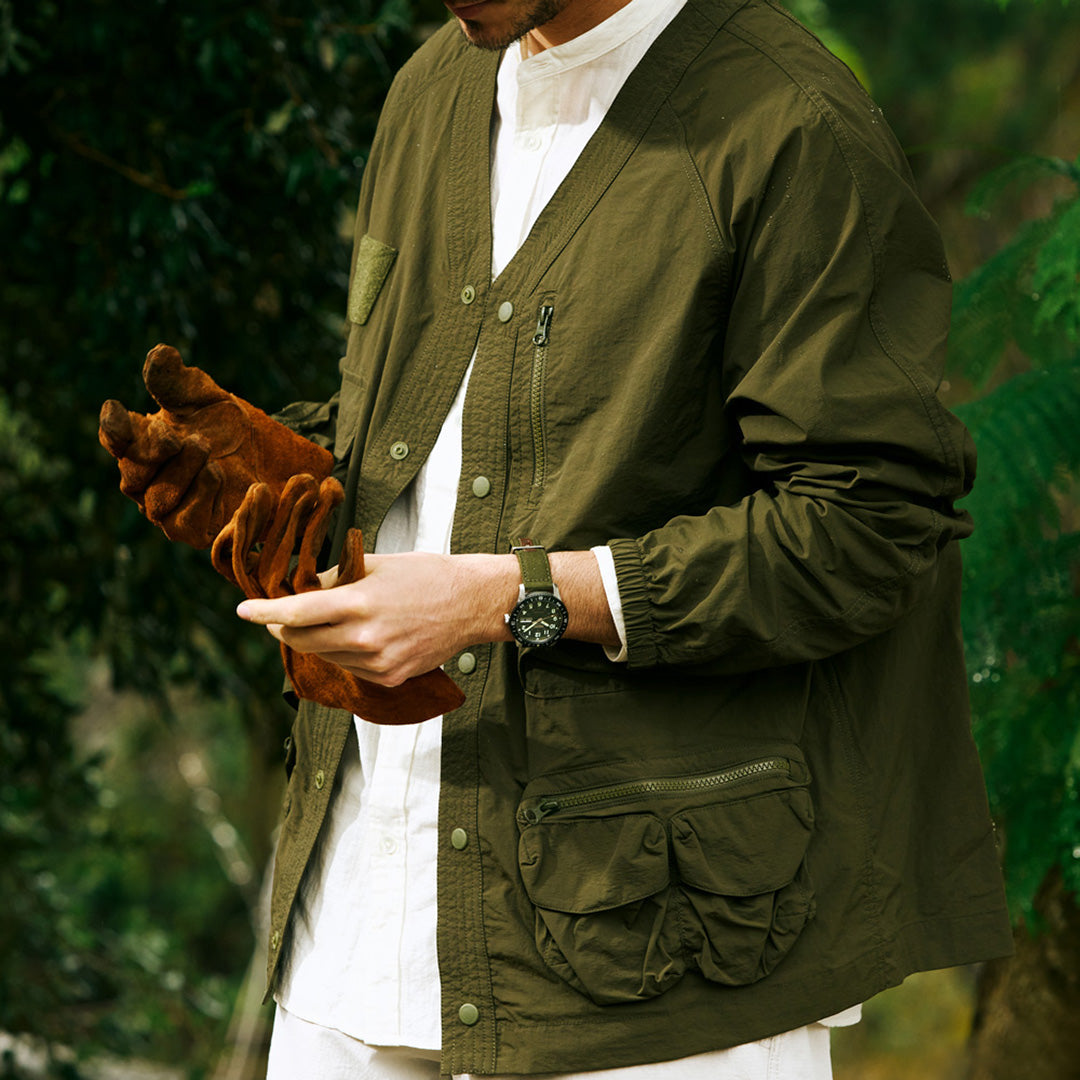An individual outdoors, dressed in an olive green jacket layered over a white shirt and putting on brown gloves, wears a Seiko 5 Sports Field Series Automatic 41mm Watch by Seiko with a durable nylon strap on their wrist. Their face is not visible, and lush greenery fills the background.