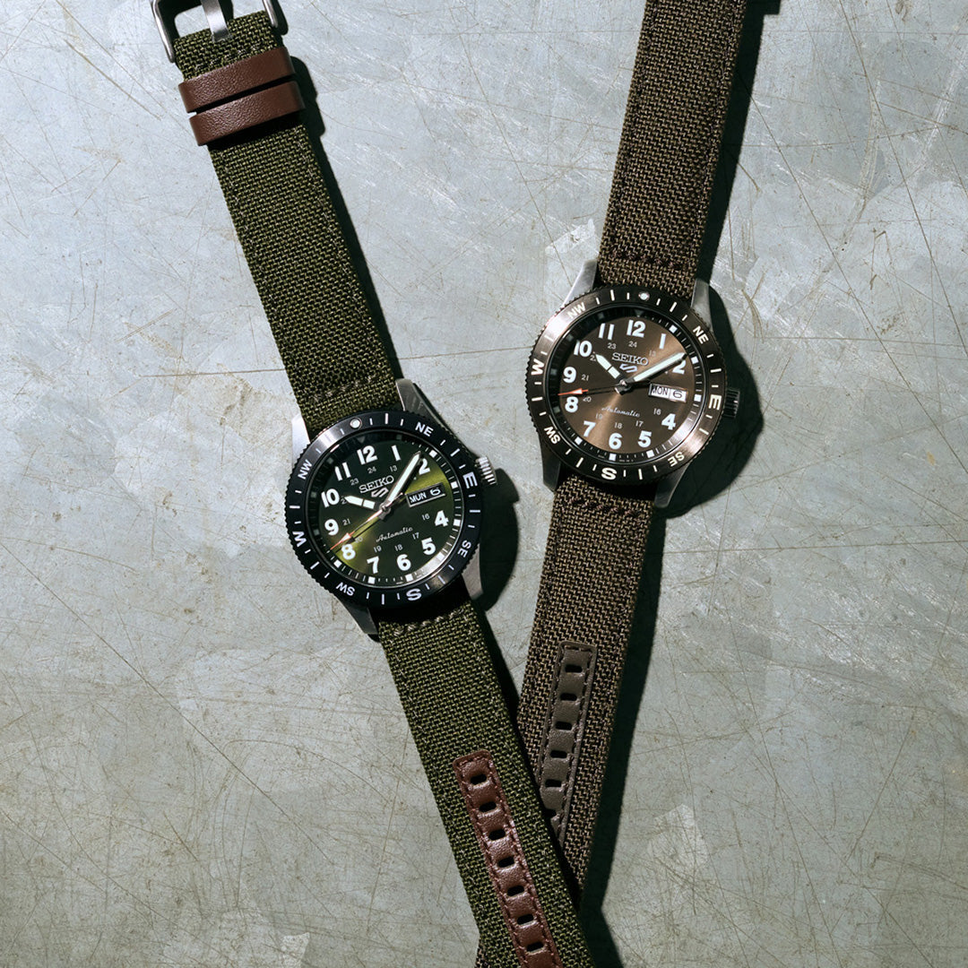 Two Seiko wristwatches with rugged nylon straps are displayed on a textured gray surface. Both feature dark faces with white numerals and indices enhanced by LumiBrite for excellent visibility in low light, as well as metallic bezels. The left watch is the Seiko 5 Sports Field Series Automatic 41mm Watch, distinguished by a brown leather accent on its strap.
