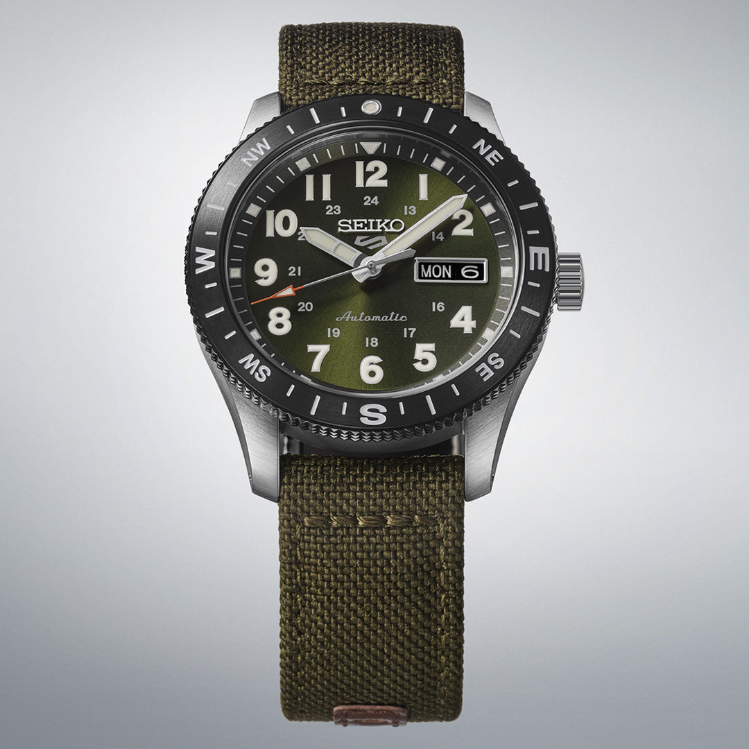 The Seiko 5 Sports Field Series Automatic 41mm Watch by Seiko features a green dial, silver bezel, and durable nylon strap. It displays hours, minutes, seconds, and day-date windows with LumiBrite accents for enhanced visibility. The background is light gray.