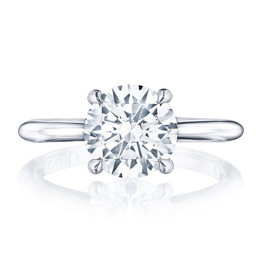 A close-up of the Tacori Founders Collection 18K White Gold Engagement Ring features a round brilliant solitaire center stone set in a four-prong setting. Crafted with premium 18K white gold, the band showcases diamond accents and has a simple, polished finish that subtly reflects light. The diamond's facets are clearly visible, adding brilliance to this masterpiece by Tacori gemologists.