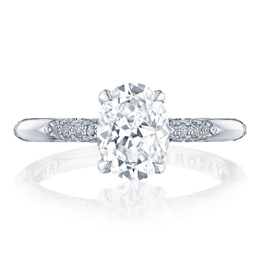 A close-up of the Tacori Founders Collection engagement ring showcases an elegant oval diamond centered on a sleek, reflective 18K white gold band. The band's design is adorned with G color diamonds and features the iconic Tacori crescent signature, enhancing its overall sparkle.