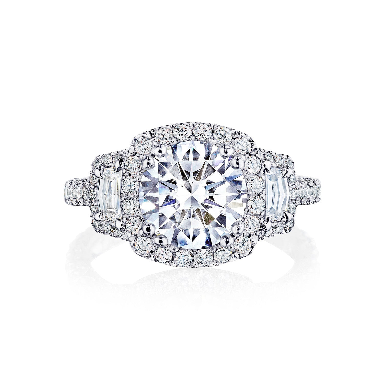 The Tacori Petite Crescent Royal T Platinum Round Brilliant Three Stone Halo Semi Mount with Diamond Side Stone Engagement Ring is meticulously crafted by Tacori gemologists. Featuring a large round center stone encircled by a halo of smaller diamonds, the band also includes additional diamonds and a geometric motif, adding to its intricate design. The ring is showcased against a white background.