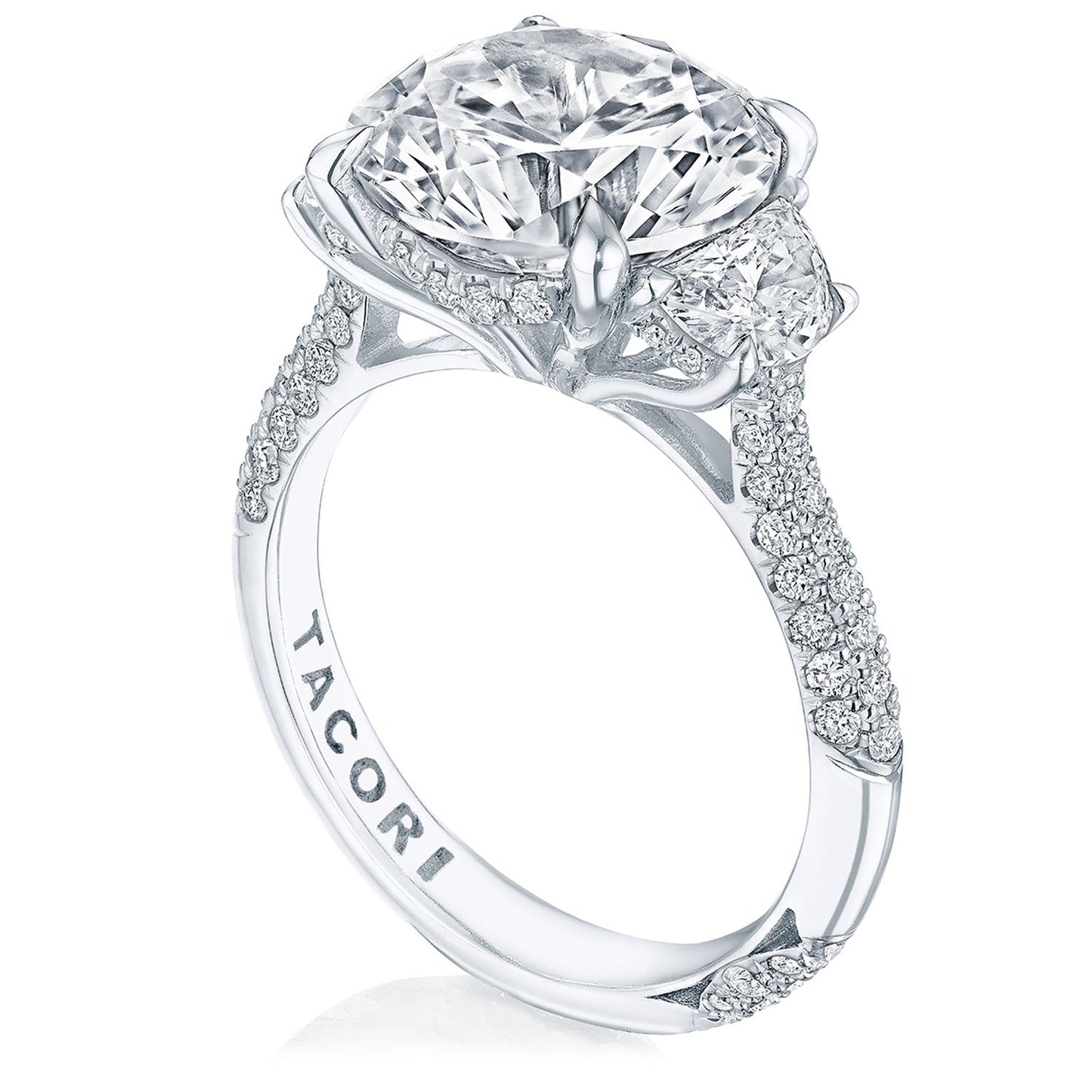 A close-up of the Tacori Founder's Collection RoyalT Platinum Round Brilliant Three Stone Semi Mount with Diamond Side Stone Engagement Ring showcases an ornate, sparkling design featuring a large oval diamond at its center. The band dazzles with pavé detailing and numerous smaller diamonds, while half-moon diamonds accentuate the sides. "TACORI" is elegantly engraved on the inside of the band.