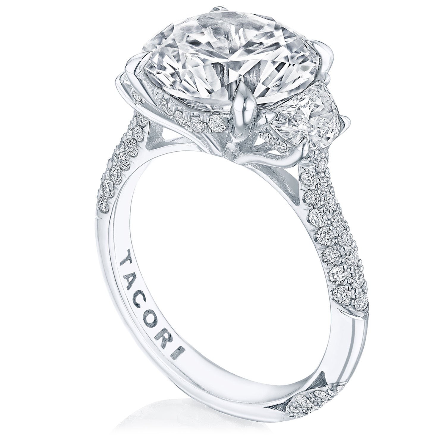 A close-up of the Tacori Founder's Collection RoyalT Platinum Round Brilliant Three Stone Semi Mount with Diamond Side Stone Engagement Ring showcases an ornate, sparkling design featuring a large oval diamond at its center. The band dazzles with pavé detailing and numerous smaller diamonds, while half-moon diamonds accentuate the sides. "TACORI" is elegantly engraved on the inside of the band.
