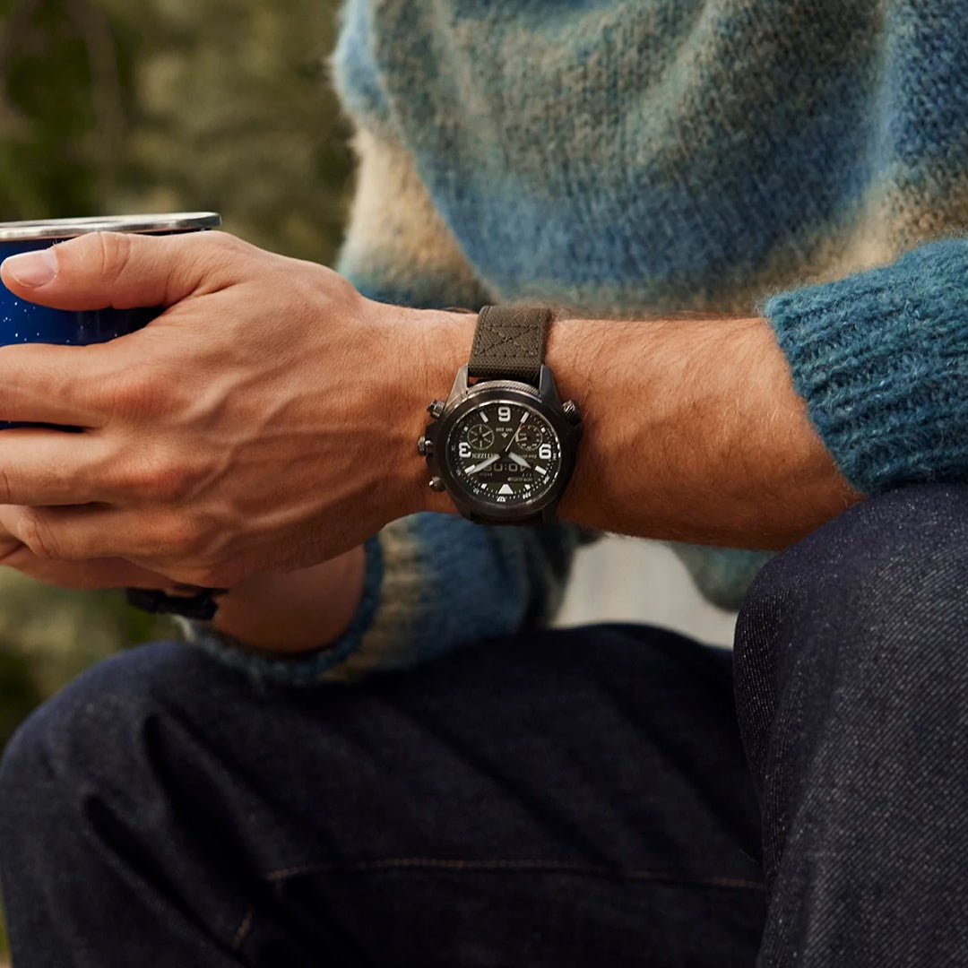 A person in a cozy, striped sweater and dark jeans holds a blue mug, wearing the CITIZEN Promaster Land U822 Eco-Drive 43.9mm Watch with a dark strap. The scene hints at a relaxed outdoor or casual setting.