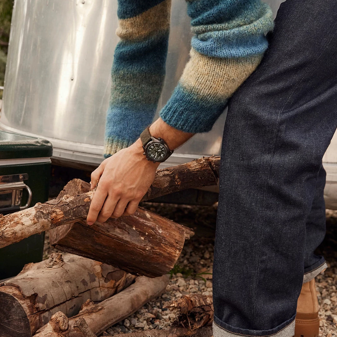 A person in a colorful striped sweater and dark jeans is picking up a log by a shiny metallic surface, wearing a CITIZEN Promaster Land U822 Eco-Drive 43.9mm watch, ready for adventure, and standing on gravel.