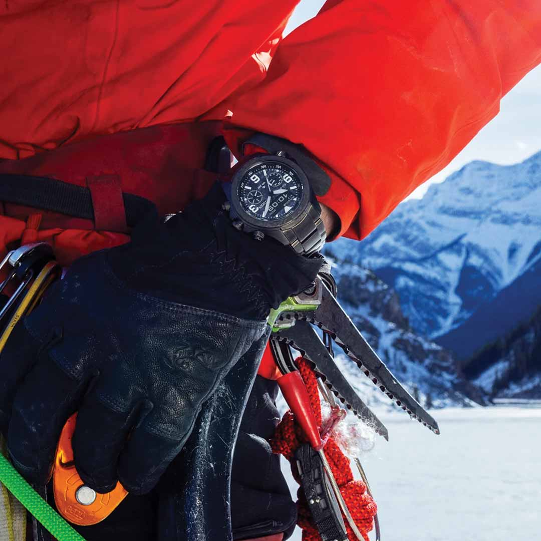 A close-up of a mountaineer wearing a red jacket and black gloves, gripping climbing gear, showcases their CITIZEN Promaster Land Limited Edition Eco-Drive 43.9mm Watch. Snow-covered mountains and a clear sky in the background amplify the spirit of exploration.