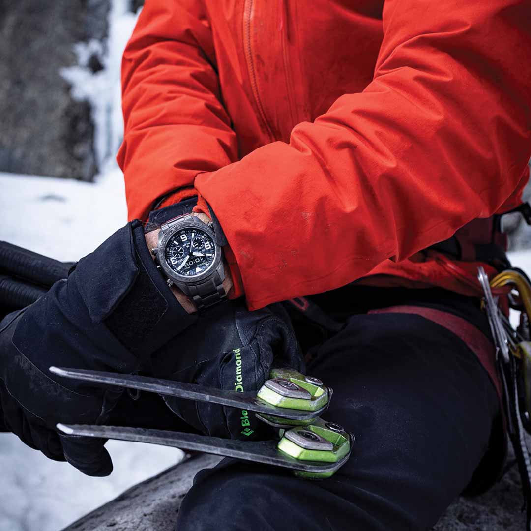 A person wearing a bright orange jacket and black gloves is holding climbing equipment, with a CITIZEN Promaster Land Limited Edition Eco-Drive 43.9mm watch on their left wrist. This rugged timepiece from CITIZEN stands out as they brave the snowy terrain in the background.