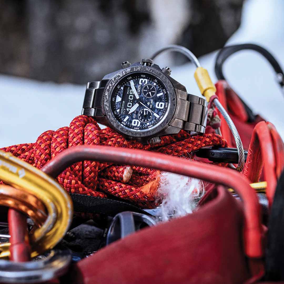 A CITIZEN Promaster Land Limited Edition Eco-Drive 43.9mm Watch with a stainless steel band is placed on top of red climbing ropes and carabiners. The snowy landscape in the background suggests a rugged, outdoor setting, capturing the adventurous spirit of the CITIZEN brand.