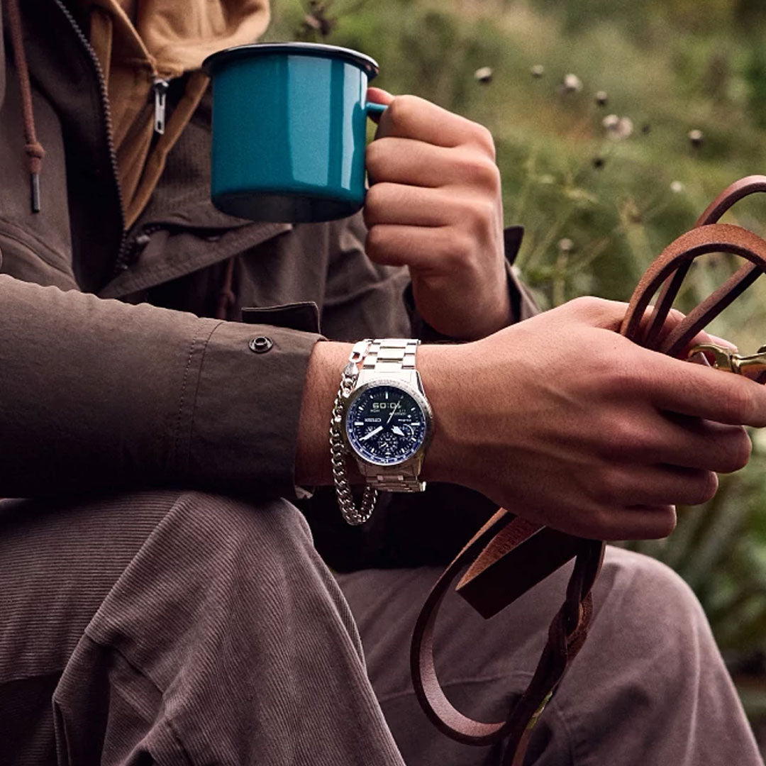 A person outdoors, surrounded by greenery, wears a CITIZEN Promaster Skyhawk U830 Eco-Drive 43mm Watch and holds leather straps in one hand and a teal mug in the other.