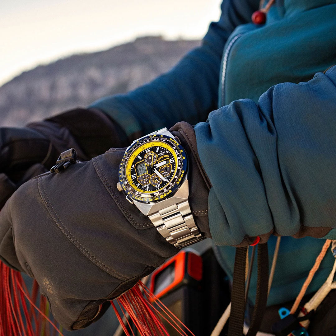 Wearing a blue jacket and gloves, a person holds red cords, showcasing the CITIZEN Promaster Skyhawk A-T Eco-Drive 46mm Watch on their wrist. A rocky landscape is blurred in the background.