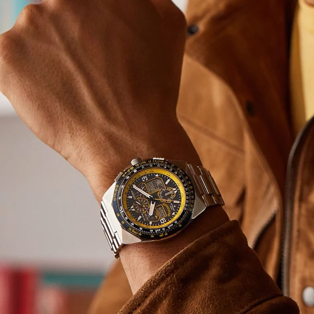 A close-up of a wrist wearing the CITIZEN Promaster Skyhawk A-T Eco-Drive 46mm Watch, featuring a silver chronograph design with black and yellow bezel, worn with a brown jacket and yellow shirt. Multiple dials are visible on the watch face.