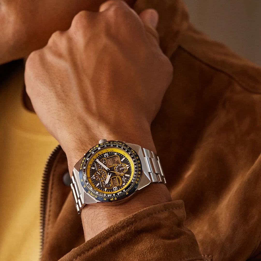 A close-up of a person’s wrist wearing the CITIZEN Promaster Skyhawk A-T Eco-Drive 46mm Watch, featuring a black and yellow bezel and intricate dial, paired with a brown suede jacket and yellow shirt.