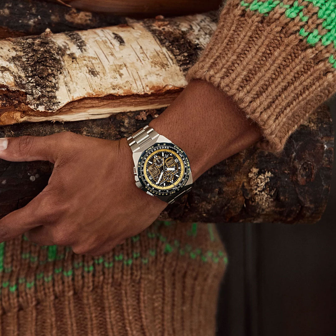 A person in a brown knit sweater with green details holds a large birch log. On their left wrist, they wear the CITIZEN Promaster Skyhawk A-T Eco-Drive 46mm Watch featuring a black and gold face.
