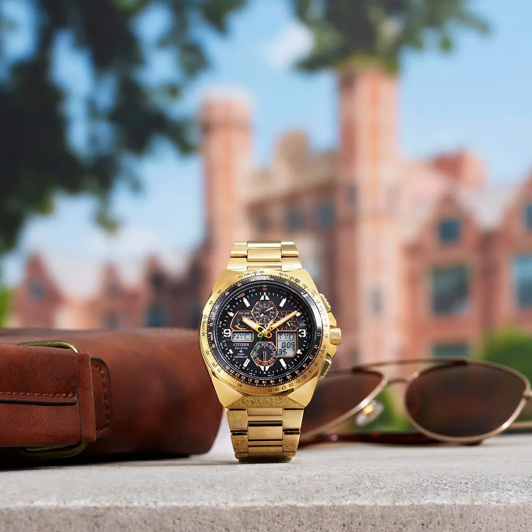 A limited edition CITIZEN Promaster Skyhawk A-T Eco-Drive 46mm watch in gold, featuring a black dial, is showcased on a stone surface alongside brown sunglasses and a leather case. The blurred backdrop of a historic building and green trees accentuates the refined elegance and precise atomic timekeeping of this exclusive CITIZEN timepiece, with only 5,200 pieces available.