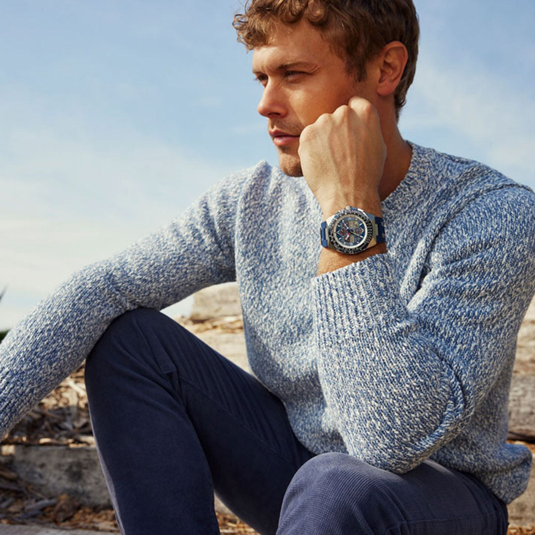 A man sits outdoors on stone steps wearing a light blue sweater and dark pants, gazing into the distance with his chin resting on his hand. On his wrist, the CITIZEN Promaster Sikorsky Eco-Drive 46mm Watch stands out against the backdrop of a clear sky.