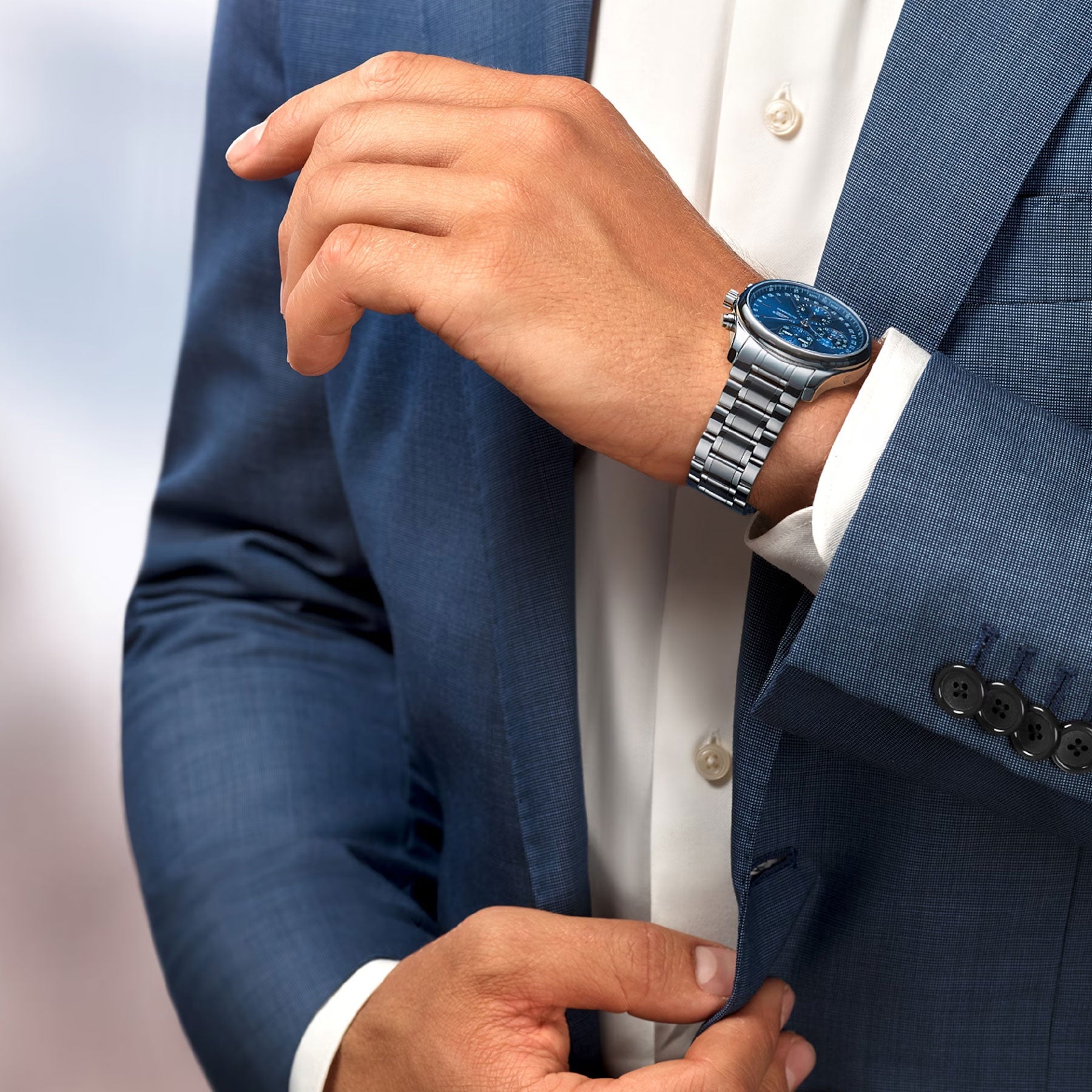 Dressed in a blue suit, an individual adjusts their cufflink, prominently displaying the distinguished Longines Master Collection Automatic 42mm Watch. The silver watch, featuring an exquisite blue face, gracefully enhances the outfit and stands out against the blurred background.