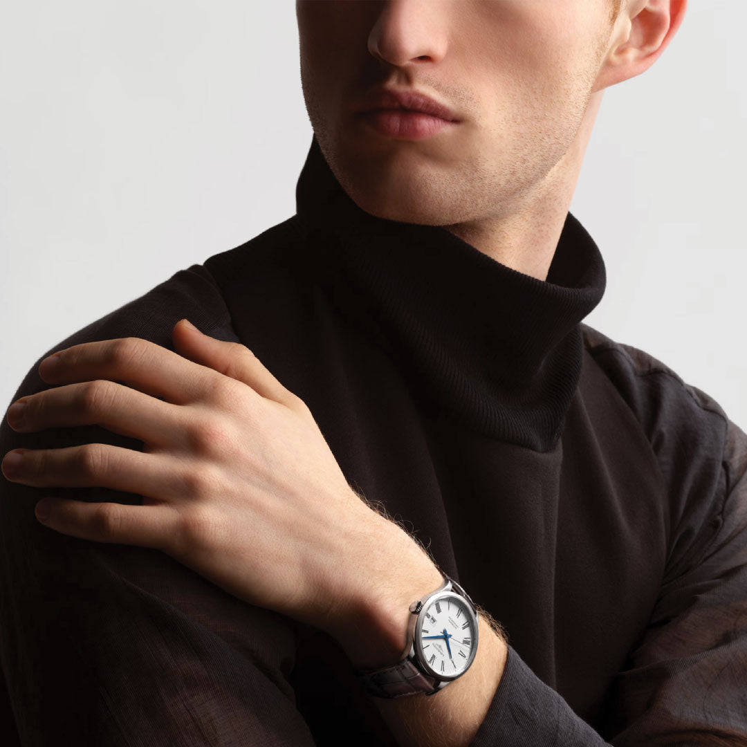 A person wearing a black turtleneck sweater and the Longines Record Automatic 40mm Watch exudes classic elegance, resting their left hand on their right shoulder. The focus is on the watch with a white face and silver strap, featuring the precise single-crystal silicon balance spring against a light gray background from Longines.