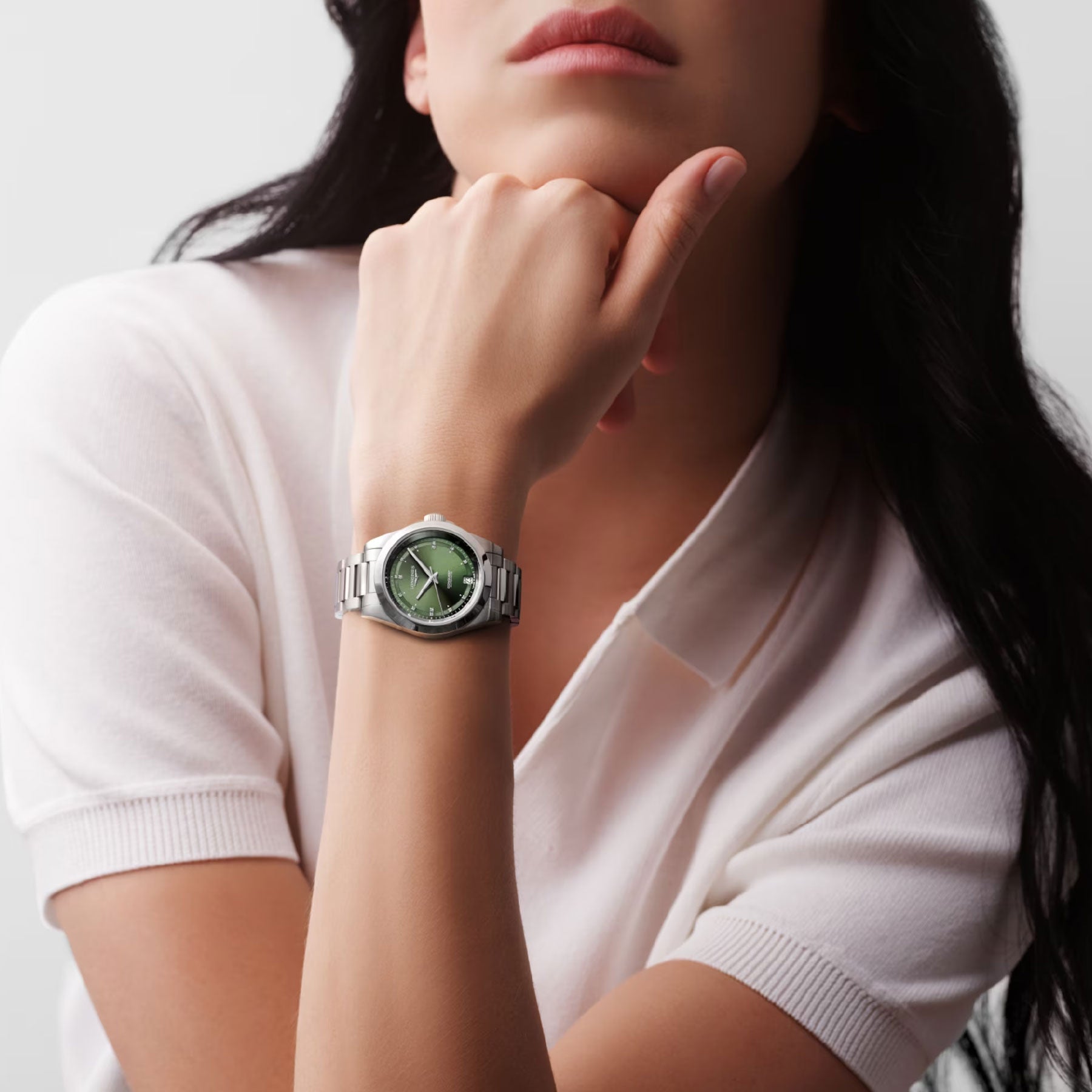 A person in a white shirt rests their chin on their hand, exuding elegance while showcasing the Longines Conquest Automatic 34mm Watch, which features a striking green face and metallic band. The background complements the scene with its soft, neutral shade.