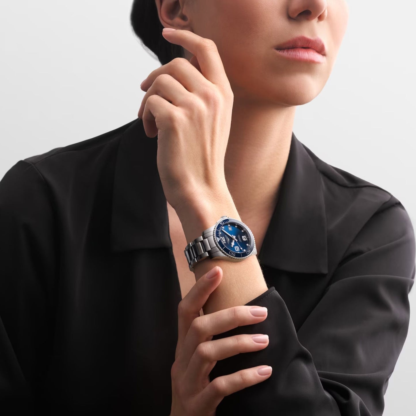 A person wearing a black shirt poses with their hand near their face, featuring a Longines Hydroconquest Automatic 39mm watch. The blue dial of this Swiss timepiece stands out against the plain white background, accentuating its elegance and detailed craftsmanship.