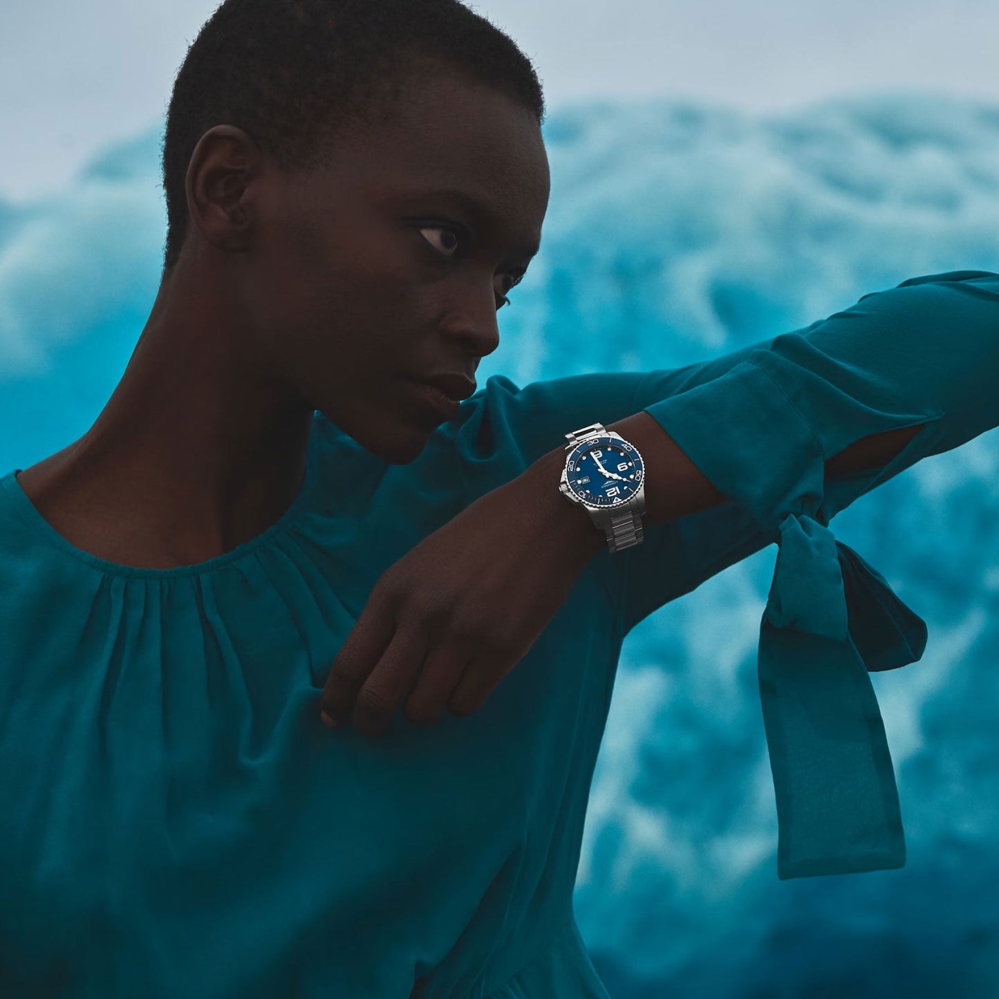 A person in a teal blouse poses with their arm raised, showcasing the stylish Longines Hydroconquest Automatic 39mm Watch. The watch features a classy blue face, and behind them is a blurred background of soft, cloud-like shapes.