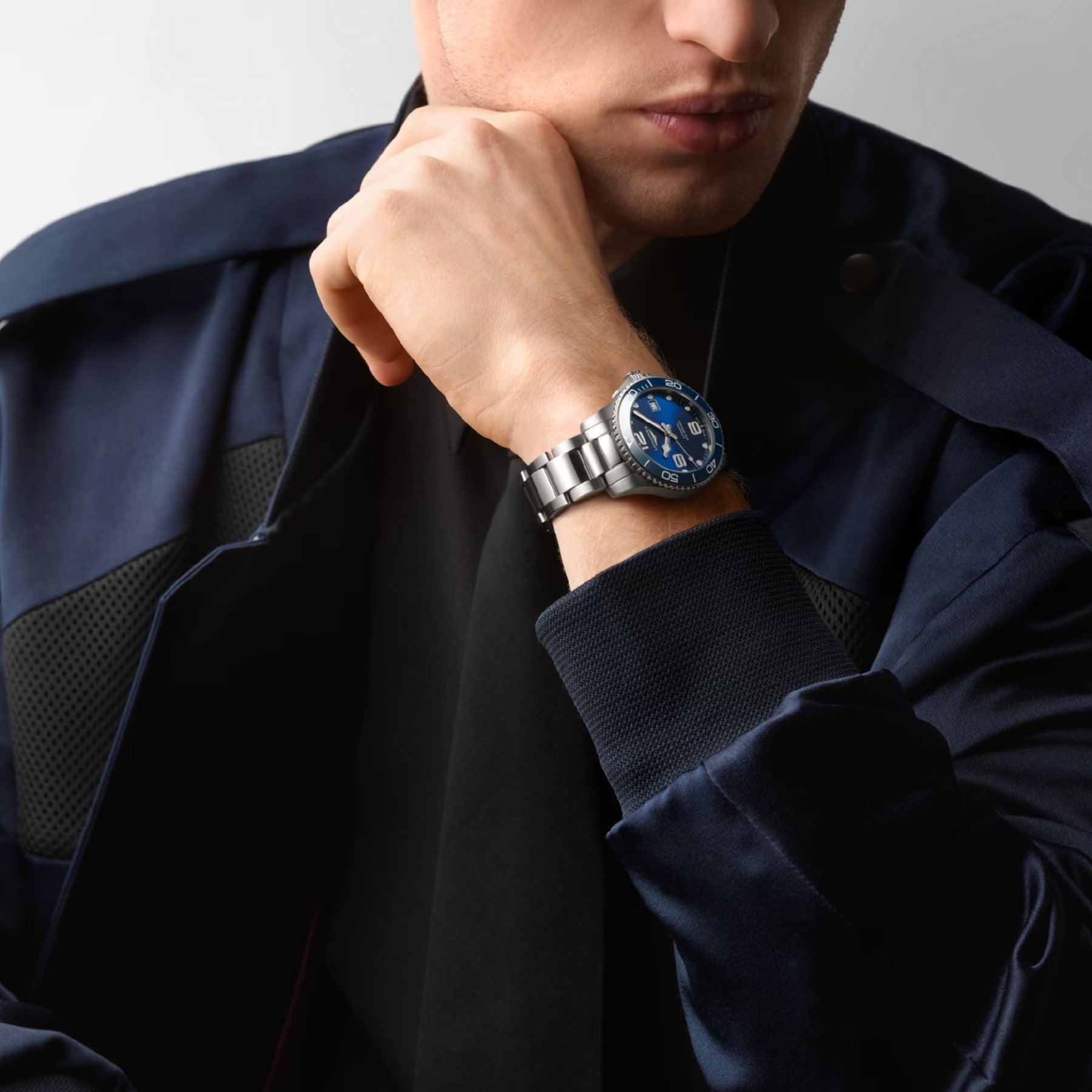 A person in a dark blue jacket and a wristwatch featuring a blue face and silver band rests their chin on their hand. This Swiss timepiece, potentially the Longines HydroConquest Automatic 39mm Watch, showcases its detailed features prominently.