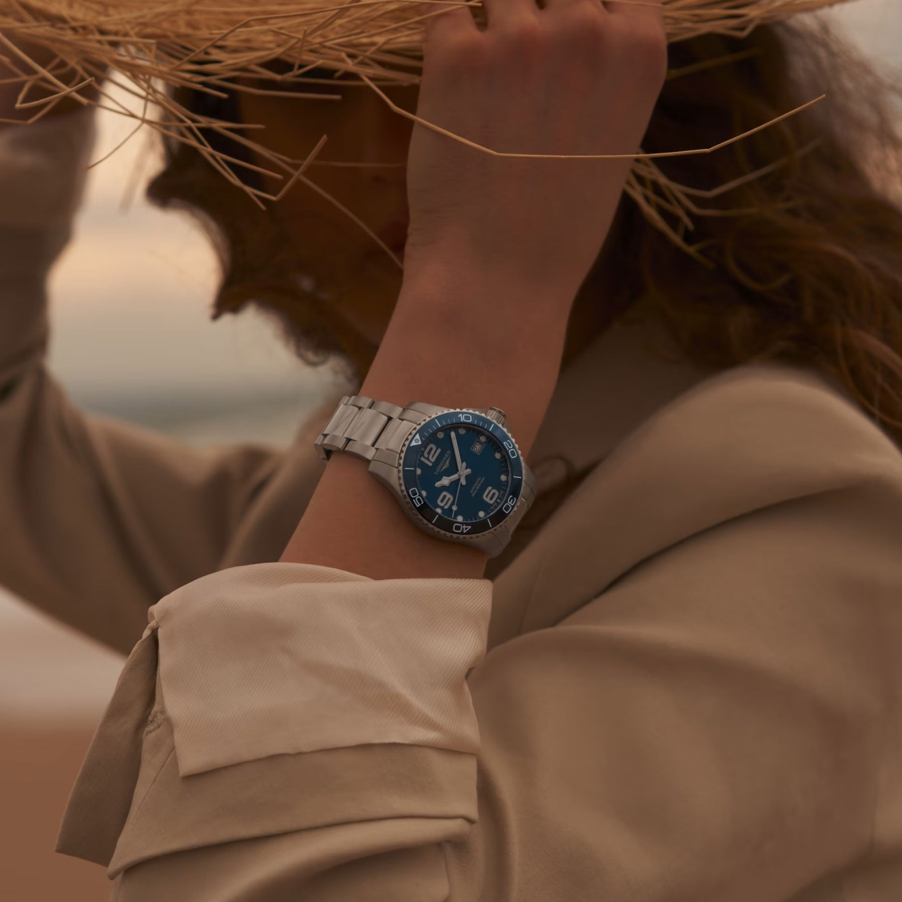 A person wearing a beige coat holds a wide-brimmed hat, revealing a Longines Hydroconquest Automatic 39mm Watch with a silver case and blue face. The background is a blurred outdoor setting, suggesting a beach or coastal location.