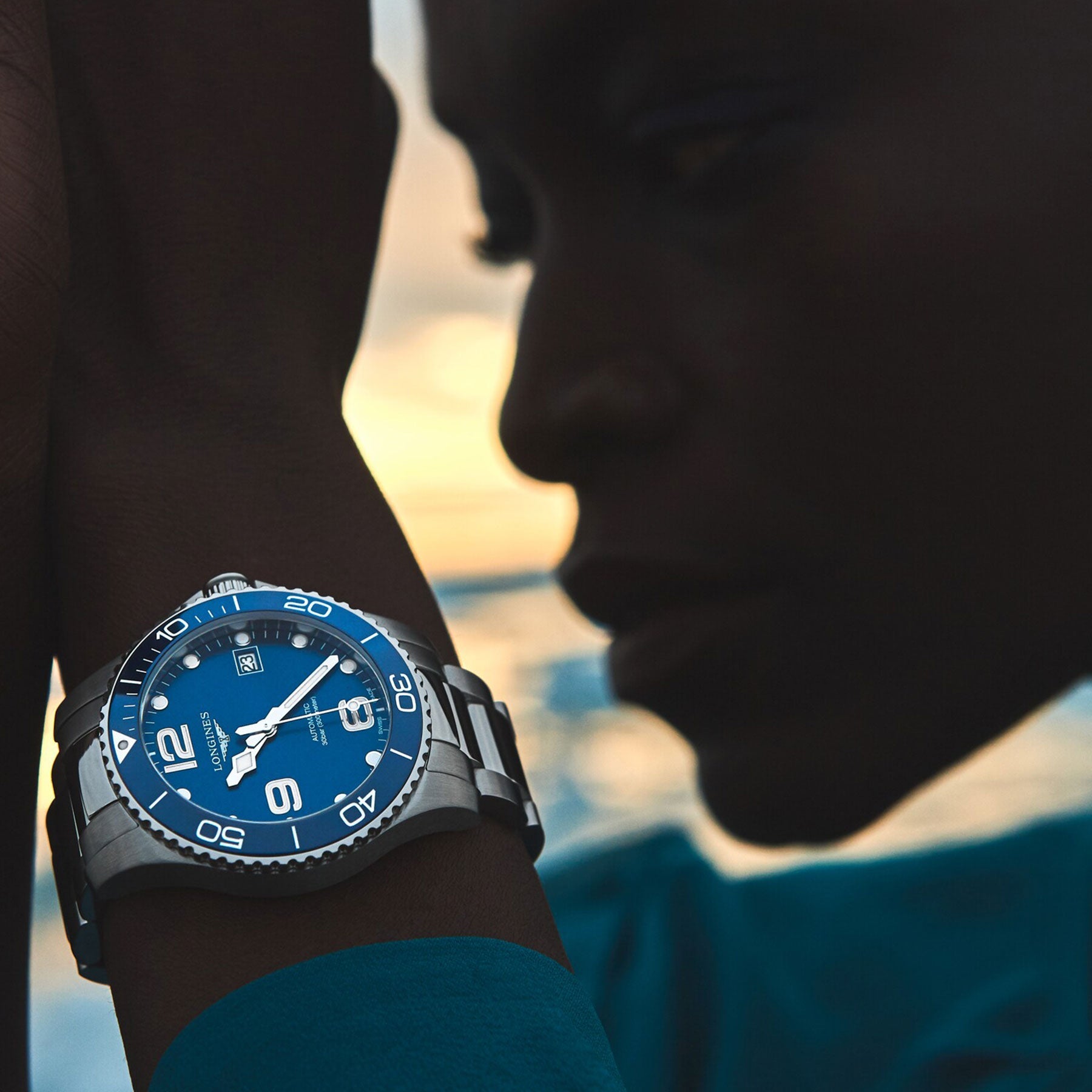 A person wearing a Longines Hydroconquest Automatic 39mm watch, featuring a silver and blue face with numeric markers and a date display. The softly blurred background suggests the warm hues of a sunset or sunrise, partially casting shadows over the wearer.